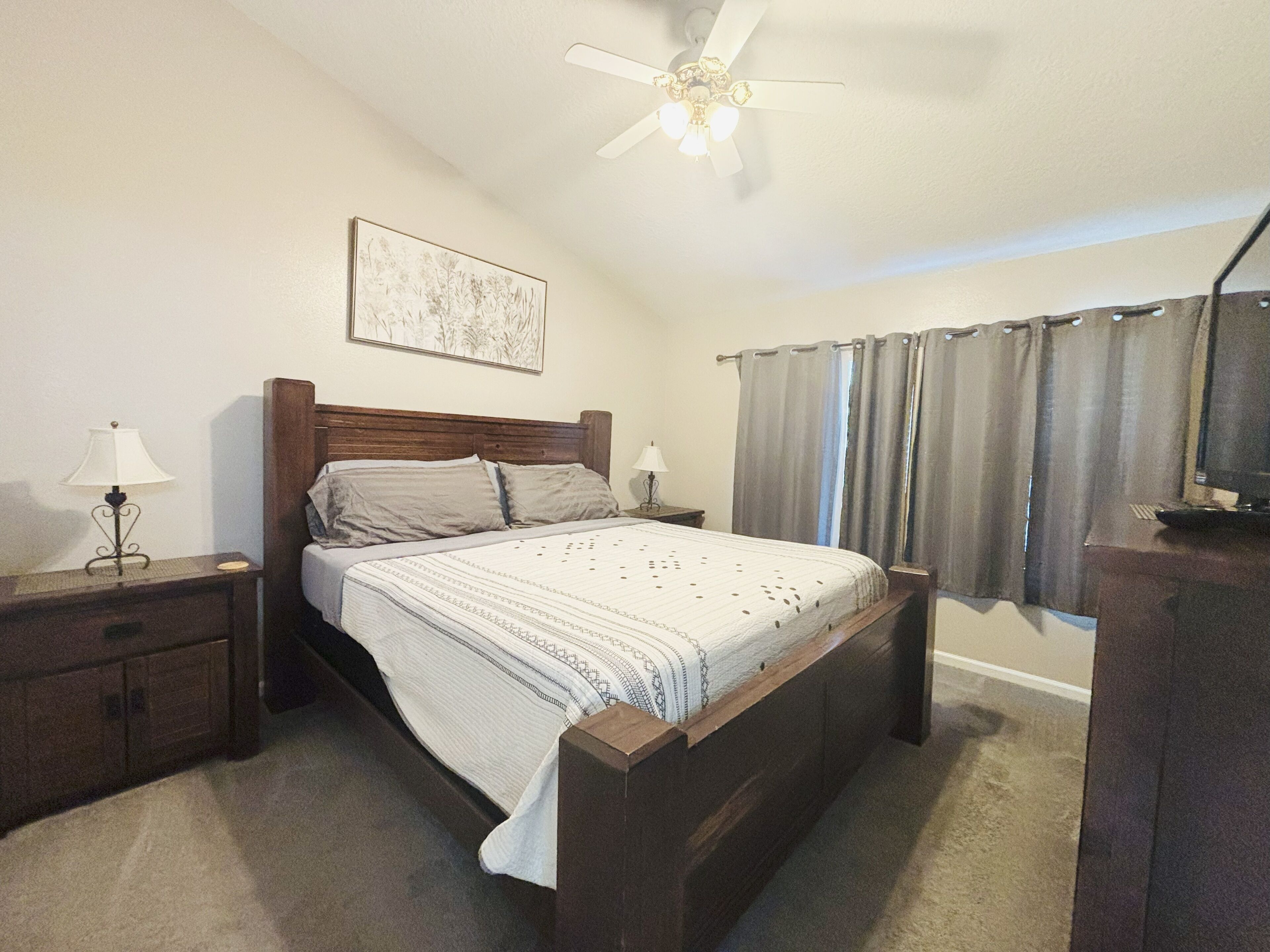 Un dormitorio con una cama de madera, dos mesitas de noche, un ventilador de techo, una ventana con cortinas y una obra de arte colgada en la pared.