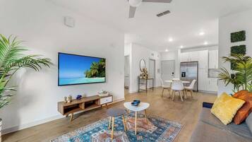 Ein modernes Wohnzimmer mit Flachbildfernseher, Deckenventilator, Essbereich mit Tisch und Stühlen sowie einer Küche im Hintergrund.
