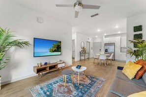 A modern living room with a flat-screen TV, a ceiling fan, a dining area with a table and chairs, and a kitchen in the background.