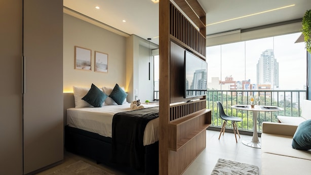 A modern hotel room with a bed, a TV, a balcony with a table and chairs, and a city view.