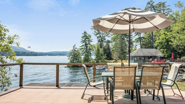 A wooden deck with a table and chairs, overlooking a lake with boats and trees. - Grand Shore Haven (Lake George)