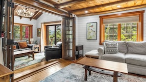 A spacious living room with wooden flooring, a fireplace, and large windows offering a view of the outdoors. - Grand Shore Haven (Lake George)