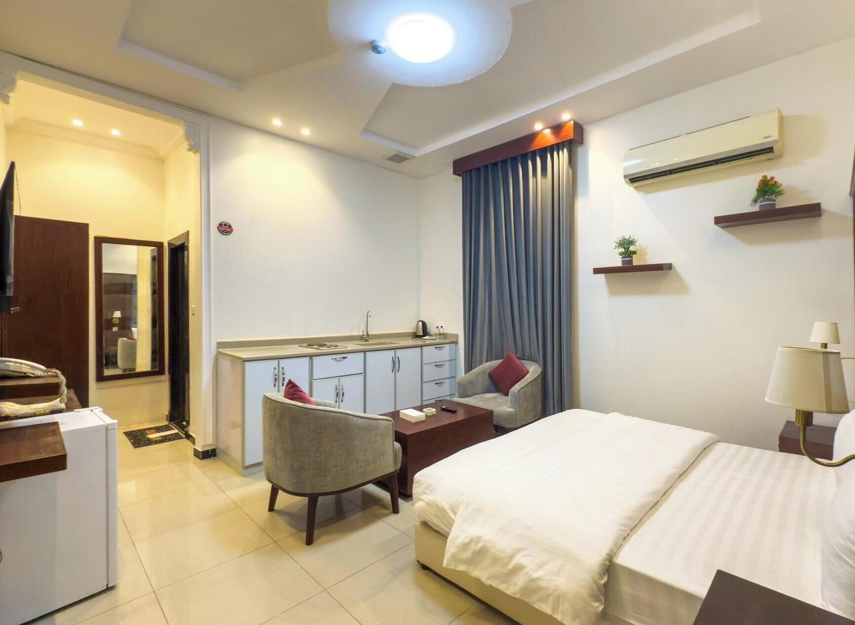 A hotel room with a bed, a desk, a chair, a small kitchenette, and a bathroom.