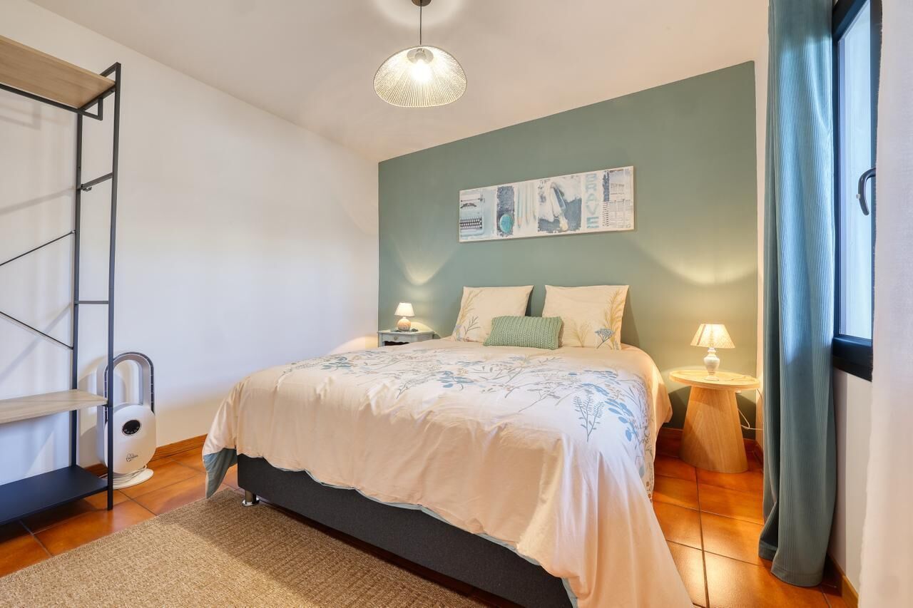 Une chambre à coucher comprenant un lit, des tables de chevet, un luminaire suspendu, une œuvre d’art accrochée au mur et une fenêtre avec des rideaux.