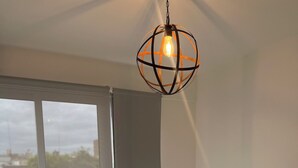 A bedroom with a bed, a potted plant, a large window with a view, and a hanging light fixture. - Esmeralda Apart (Marcos Juárez)