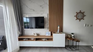 Ein modernes Wohnzimmer mit an einer Marmorwand montiertem Fernseher, einem Holzkonsolentisch mit Dekoration und einem runden Holzhock, sowie einer Schiffsraduhr.