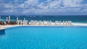 Blick auf ein Schwimmbecken mit der Beschilderung 'THE BEACH CLUB', Liegestühlen und freiem Blick auf den Ozean.