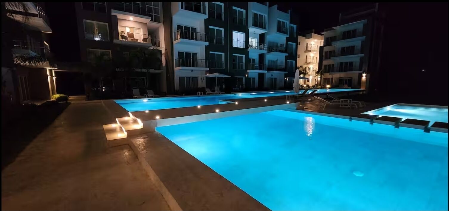 A nighttime view of a hotel pool area with multiple pools and lounge chairs, surrounded by multi-story buildings with balconies and outdoor lighting.