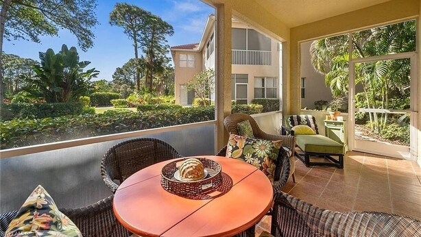 Een balkon met rieten meubilair, een ronde tafel en uitzicht op een tuin en gebouw.