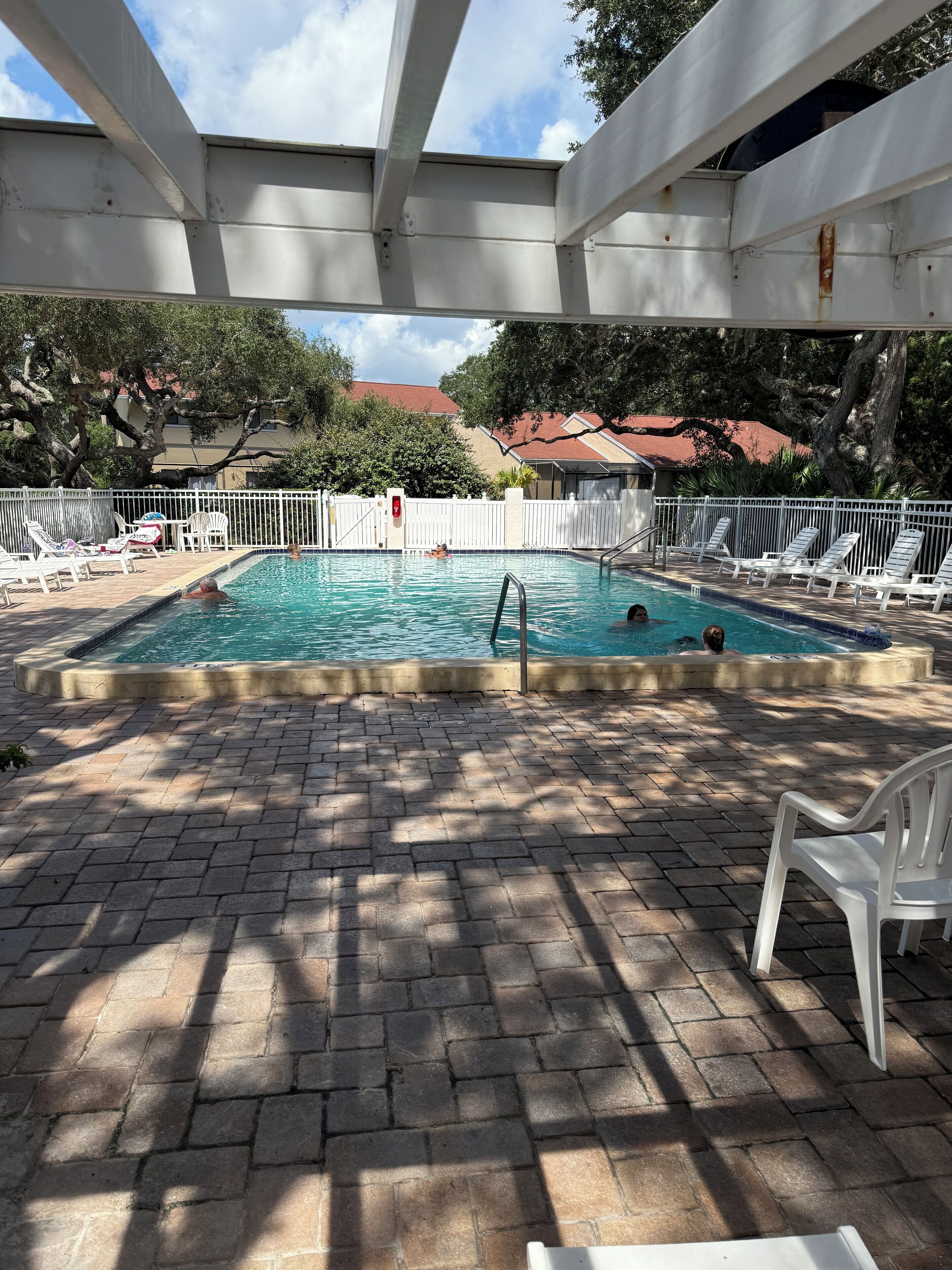 A swimming pool with a brick deck, surrounded by white lounge chairs and a white fence.