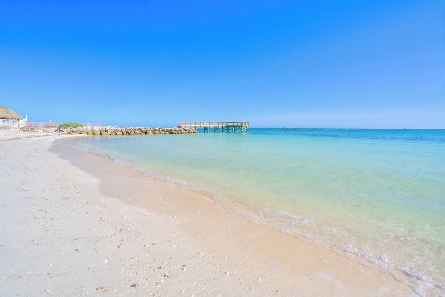 A beach with clear turquoise water, a wooden pier, and a white building in the background.