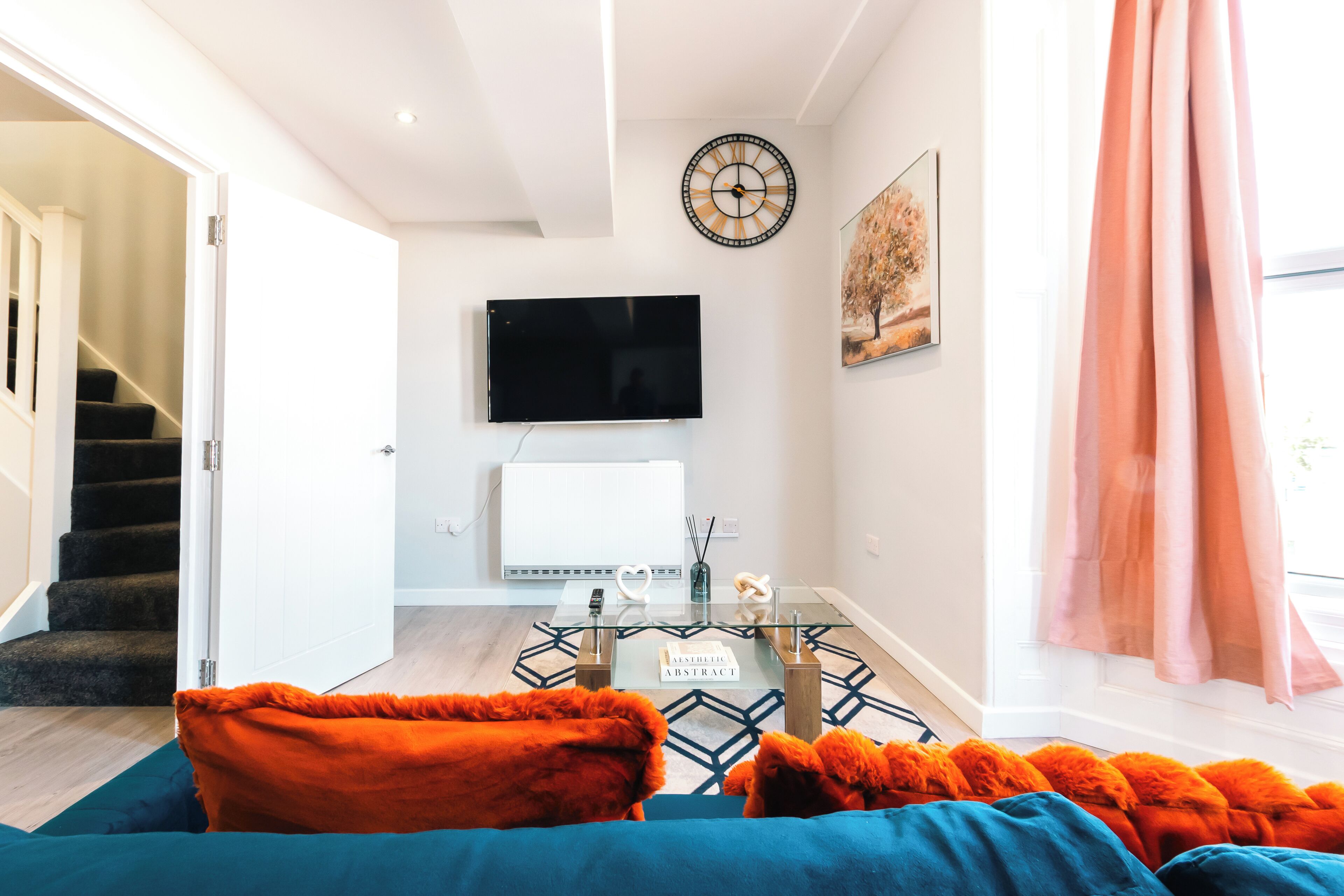 A modern living room with a flat-screen TV mounted on the wall, a glass coffee table, and a sofa with orange and blue cushions.