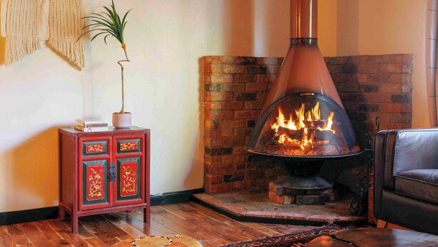 A cozy living room with a wood-burning stove, a small cabinet with decorative panels, and a potted plant. - La Forêt Retreat, Hiking, Lakes, Dog Friendly! (Running Springs)