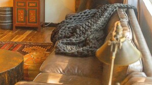 A cozy living room with a leather sofa, a knitted throw blanket, a lamp, a small cabinet, and a plant. - La Forêt Retreat, Hiking, Lakes, Dog Friendly! (Running Springs)