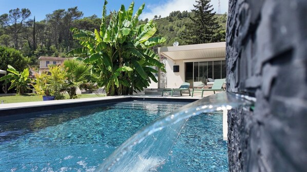 A swimming pool with a waterfall feature, surrounded by lush greenery and a modern building with a large window.