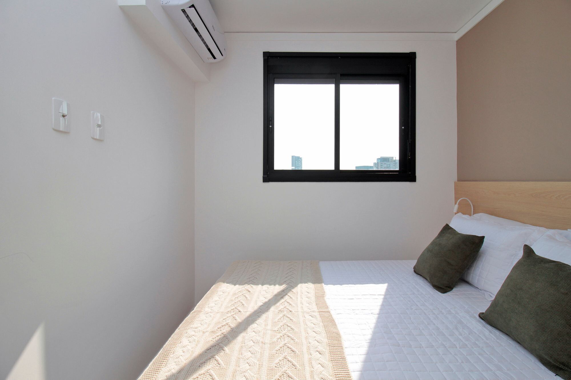 Une petite chambre bien éclairée, avec un lit simple, une fenêtre à cadre noir et un appareil de climatisation fixé au mur.
