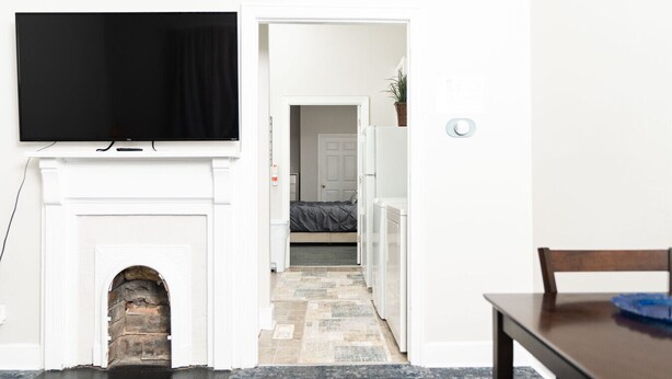 A hallway leading to a bedroom with a bed, a TV mounted above a fireplace, and a dining area with a table and chairs. - Sky cottage near downtown (Columbus)