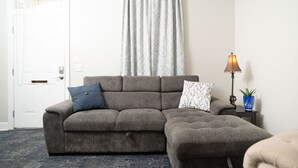A grey sectional sofa with two pillows, a lamp, and a potted plant in a living room with a patterned carpet and a white door. - Sky cottage near downtown (Columbus)