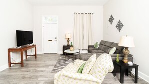 A living room with a sofa, a coffee table, a TV on a stand, and a patterned armchair. - Apple cottage near downtown (Columbus)