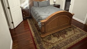 A bedroom with a wooden bed, a ceiling fan, a nightstand with a lamp, and a patterned rug. - Oak Bungalow in Midtown (Columbus)