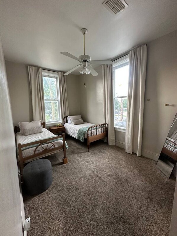 A bedroom with two single beds, a ceiling fan, large windows with curtains, and a carpeted floor.