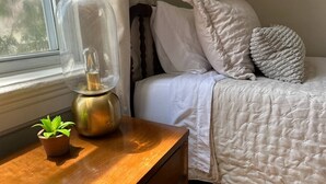 Un dormitorio con mesita de noche de madera, lámpara de latón, planta pequeña en maceta, cama con ropa de cama y almohadas blancas, y ventana con cortinas.
