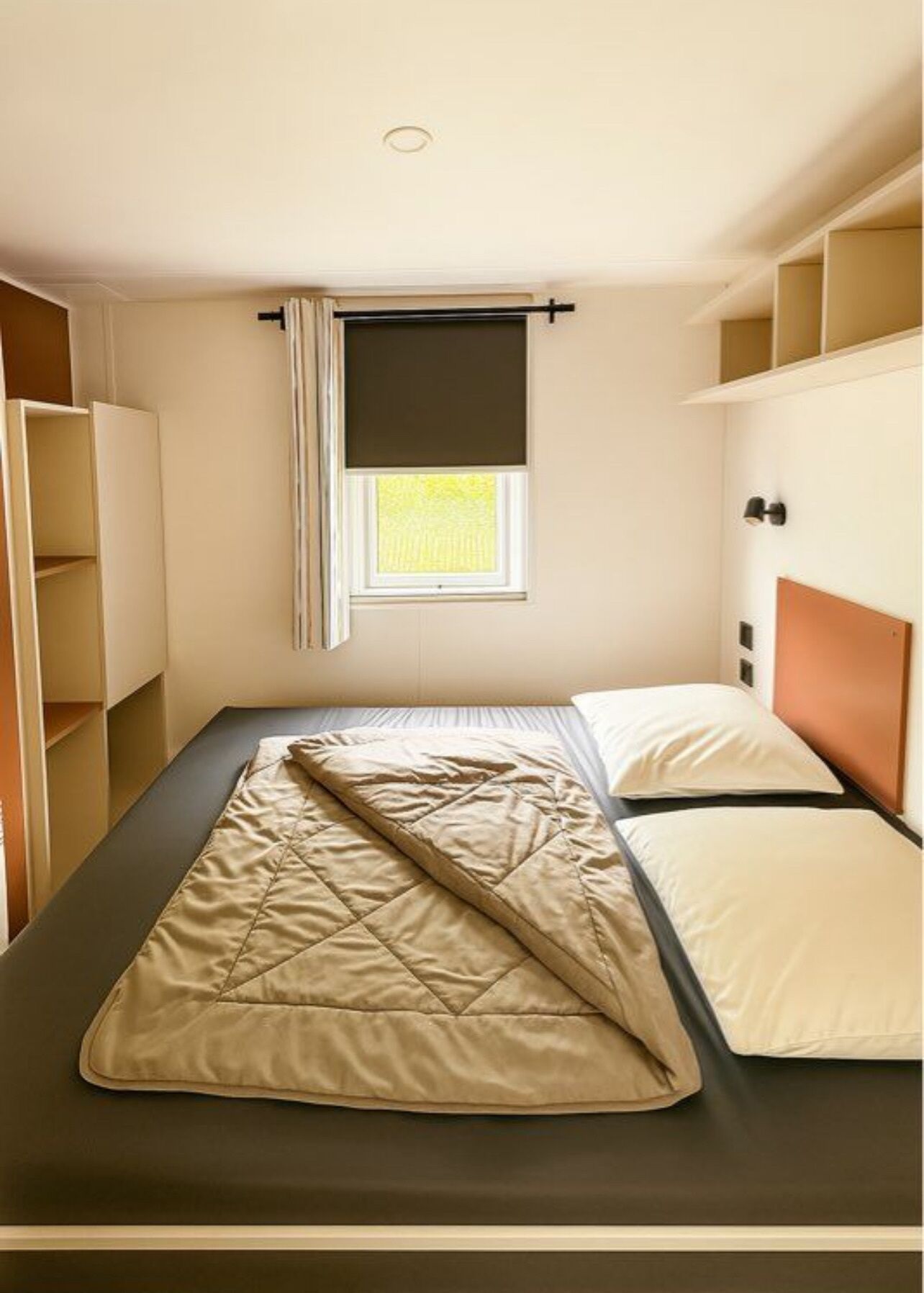 Una cama individual con colcha, una ventana con persiana y estanterías empotradas en las paredes.