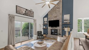 Een moderne woonkamer met een stenen openhaard, een grote flatscreen-tv en een balkon met uitzicht op groen.