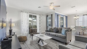 A modern living room with a grey sofa, a white coffee table, and a patterned ottoman. There is a ceiling fan, a dining area with a table and chairs, and large windows with curtains. - Kissimmee Retreat  6BR House Sleeps 14 (Kissimmee)