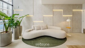 A modern interior with a curved white sofa, potted plants, and a floor lamp. - Studio With Barbecue 180m From the Beach - Sfr0302 (Bombinhas)