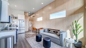 Eine moderne Küche mit integrierten Geräten, ein Essbereich mit Tisch und Stühlen, ein graues Sofa mit Kissen und ein großes Fenster, das für natürliches Licht sorgt.
