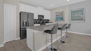 A modern kitchen with white cabinets, a stainless steel refrigerator, a microwave, and a coffee machine. There are three bar stools and a large window with a painting.