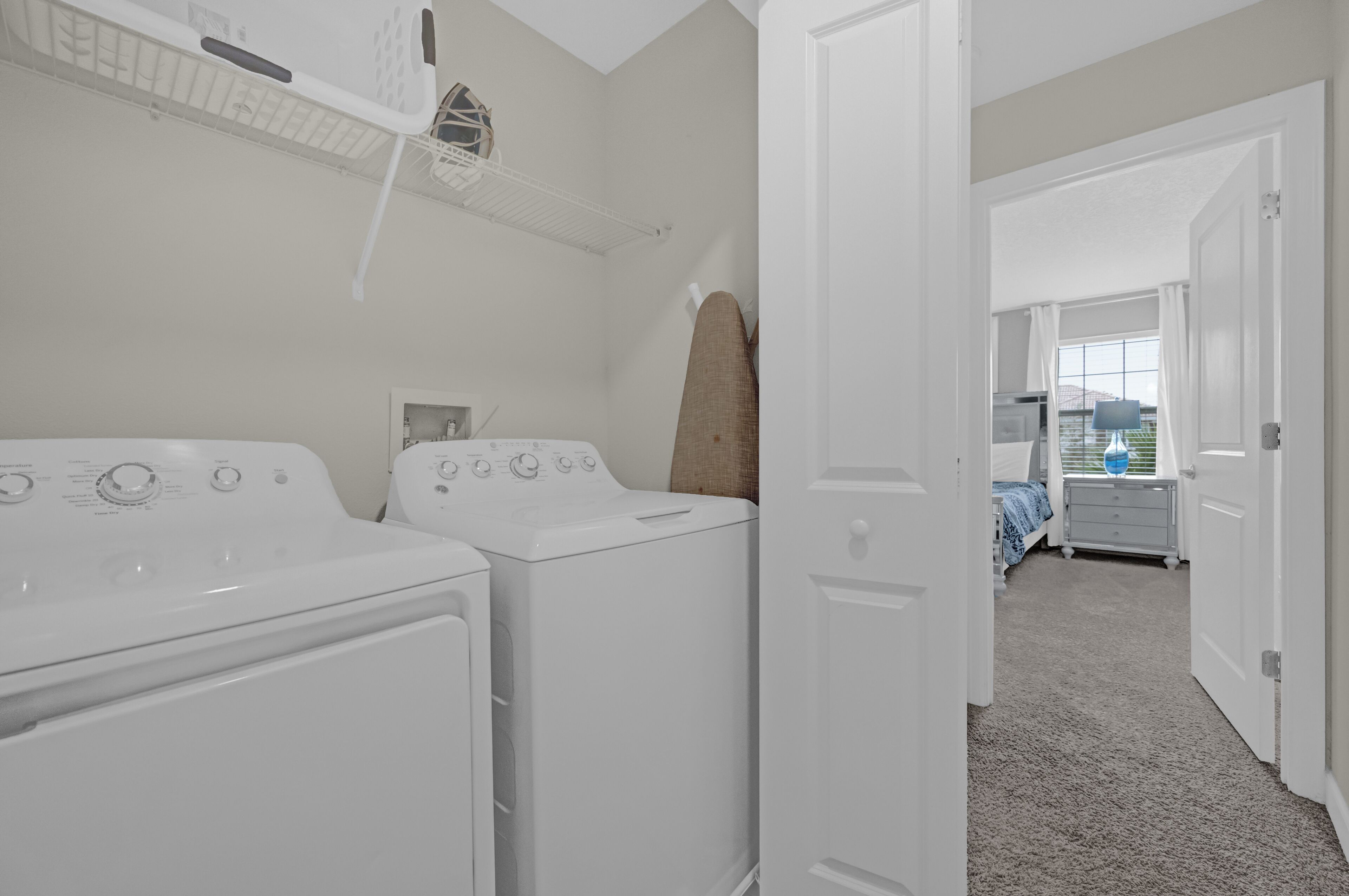 A laundry area with two white washers and dryers, a drying rack, and a door leading to a bedroom with a bed and a nightstand.