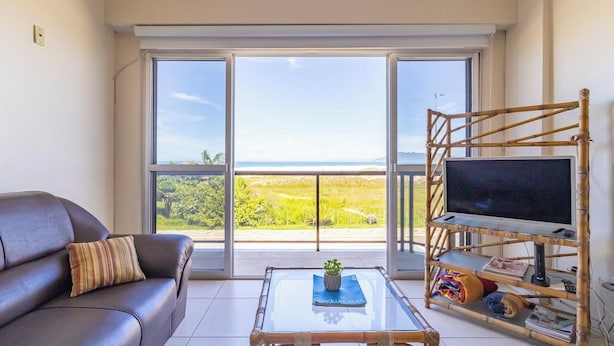 A living room with a sofa, a coffee table with a magazine, a television on a bamboo stand, and a large window with a view of greenery and a beach.