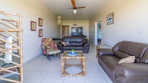 A living room with a brown leather sofa, a glass-top coffee table, a black leather couch, and a bamboo chair. There is a ceiling fan and a wall-mounted light fixture.