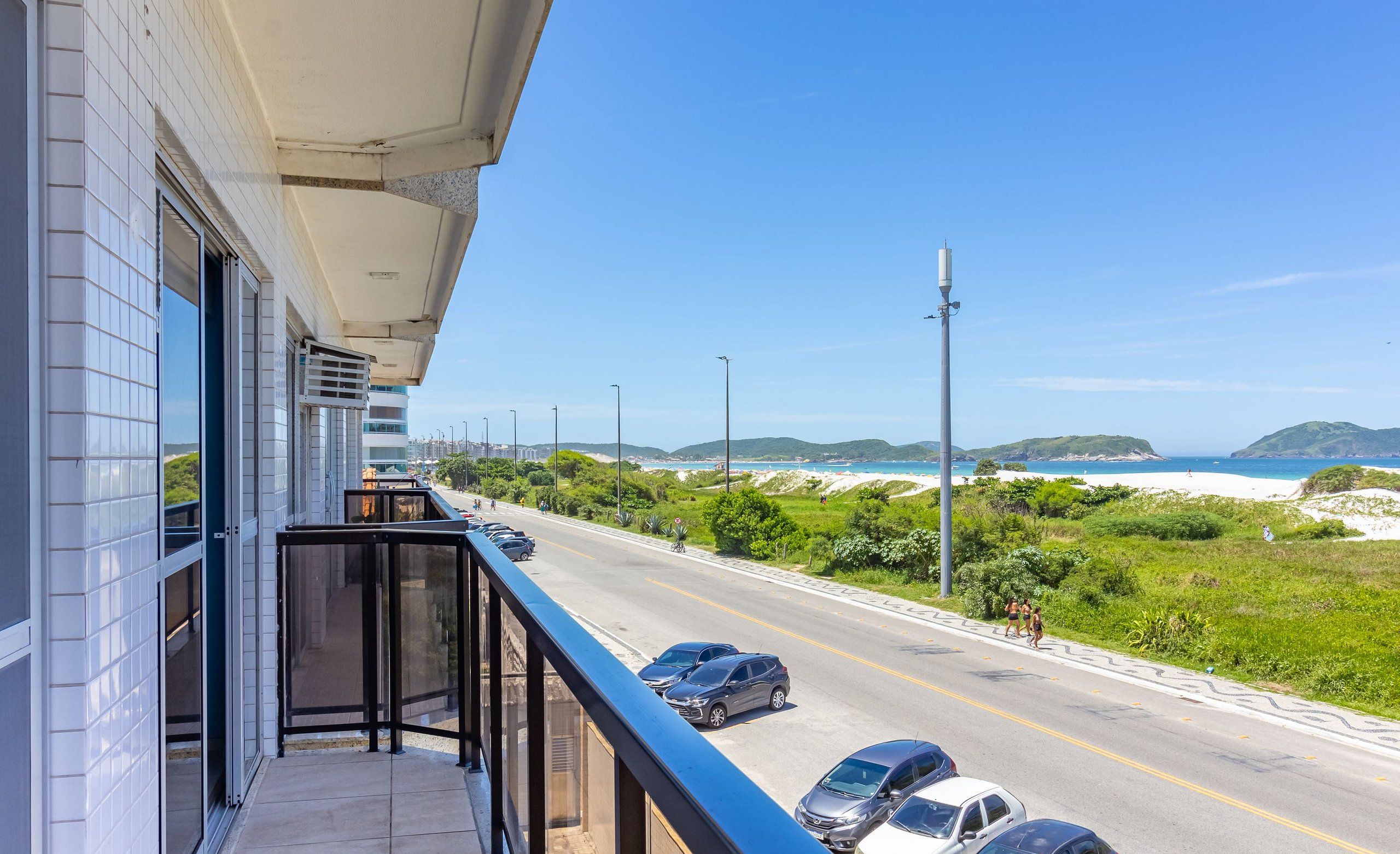 Uma varanda com vista para uma estrada, carros estacionados e uma praia ao longe.