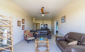 A living room with a brown leather sofa, a glass-top coffee table, a black leather couch, and a bamboo chair. There is a ceiling fan and a wall-mounted light fixture. - Apartamento 3 Quartos Cabo Frio\/RJ LDN201 (Cabo Frio)