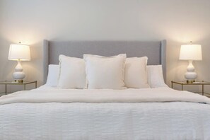 Una cama bien hecha con un cabecero gris, ropa de cama blanca y dos lámparas de noche sobre mesitas de noche doradas.