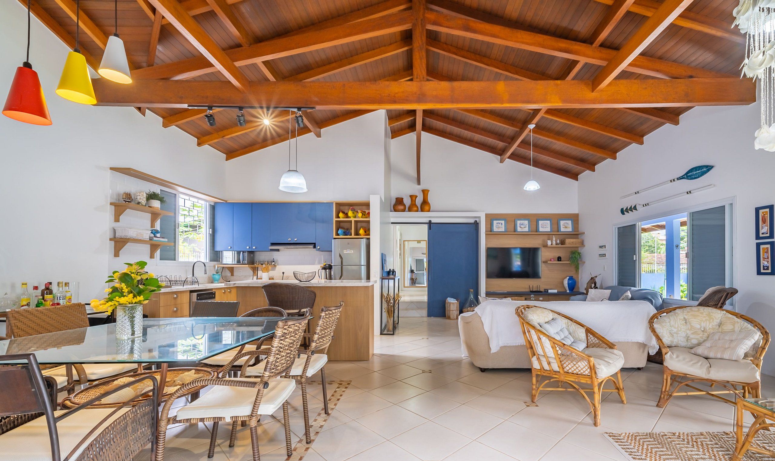 Uma sala de estar ampla com teto alto, uma mesa de jantar com cadeiras de vime, uma cozinha com armários azuis e uma televisão na sala.
