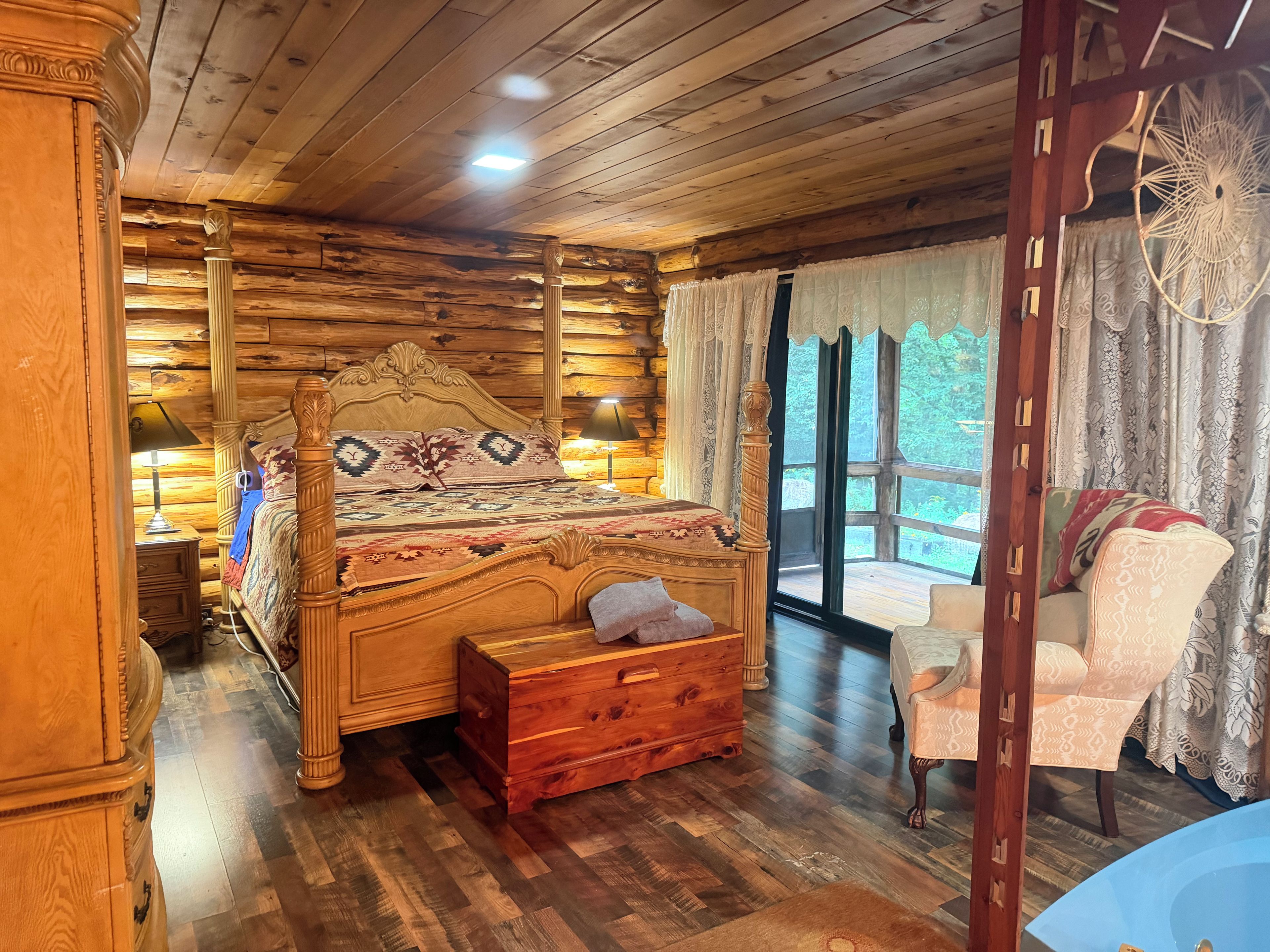 Une chambre à coucher aménagée dans une cabane en bois, avec un lit, des tables de chevet, une commode, une fenêtre avec des rideaux et une vue sur l’extérieur.