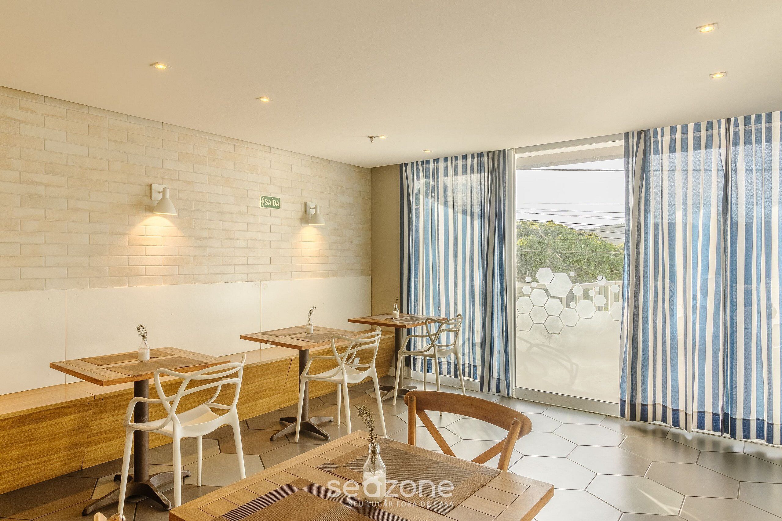 Un restaurante moderno con mesas de madera y sillas blancas, una pared de ladrillo y ventanas amplias con cortinas a rayas.