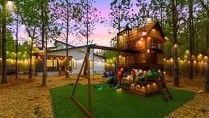 A wooden playhouse with a slide and swing set, surrounded by trees and string lights. - Kid Friendly! Foosball, Playset, 3 Arcades (Broken Bow)