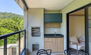 En balkon med spisebord, stole, et tekøkken med skabe og vask, samt udsigt til en frodig grøn bakke.