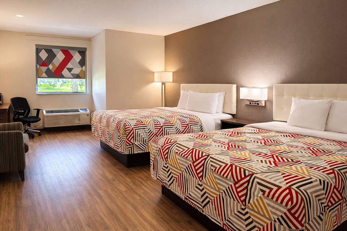 A hotel room with two beds, a desk, a chair, a window with a view, and a wall-mounted lamp.