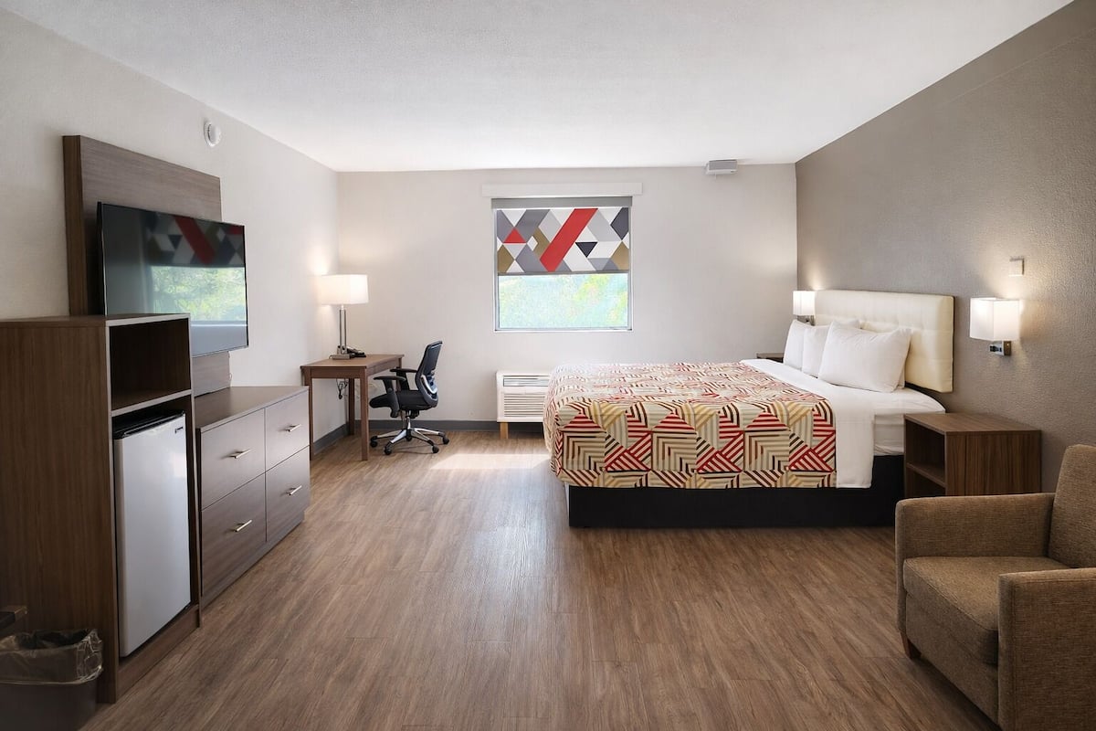 A hotel room with a bed, desk, chair, TV, and a window with a geometric pattern.