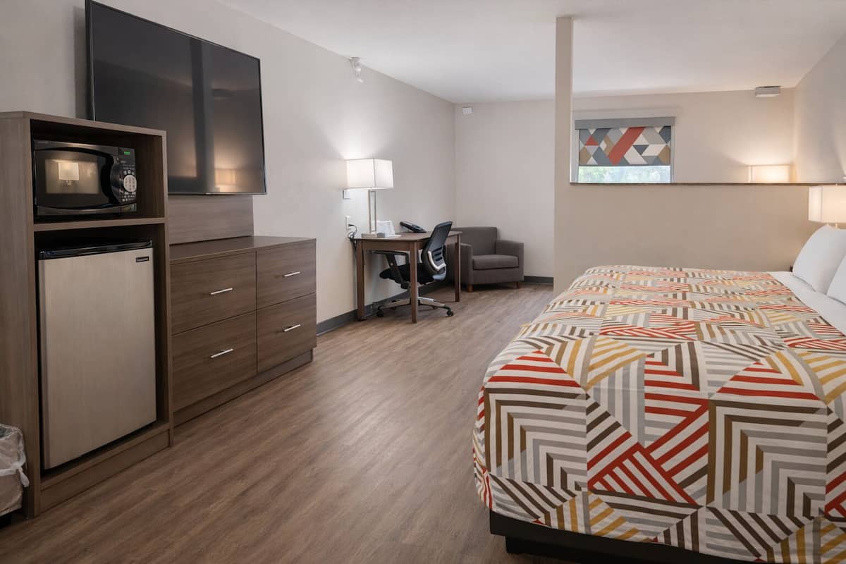 A hotel room with a bed, a desk with a chair, a microwave, a refrigerator, and a TV.
