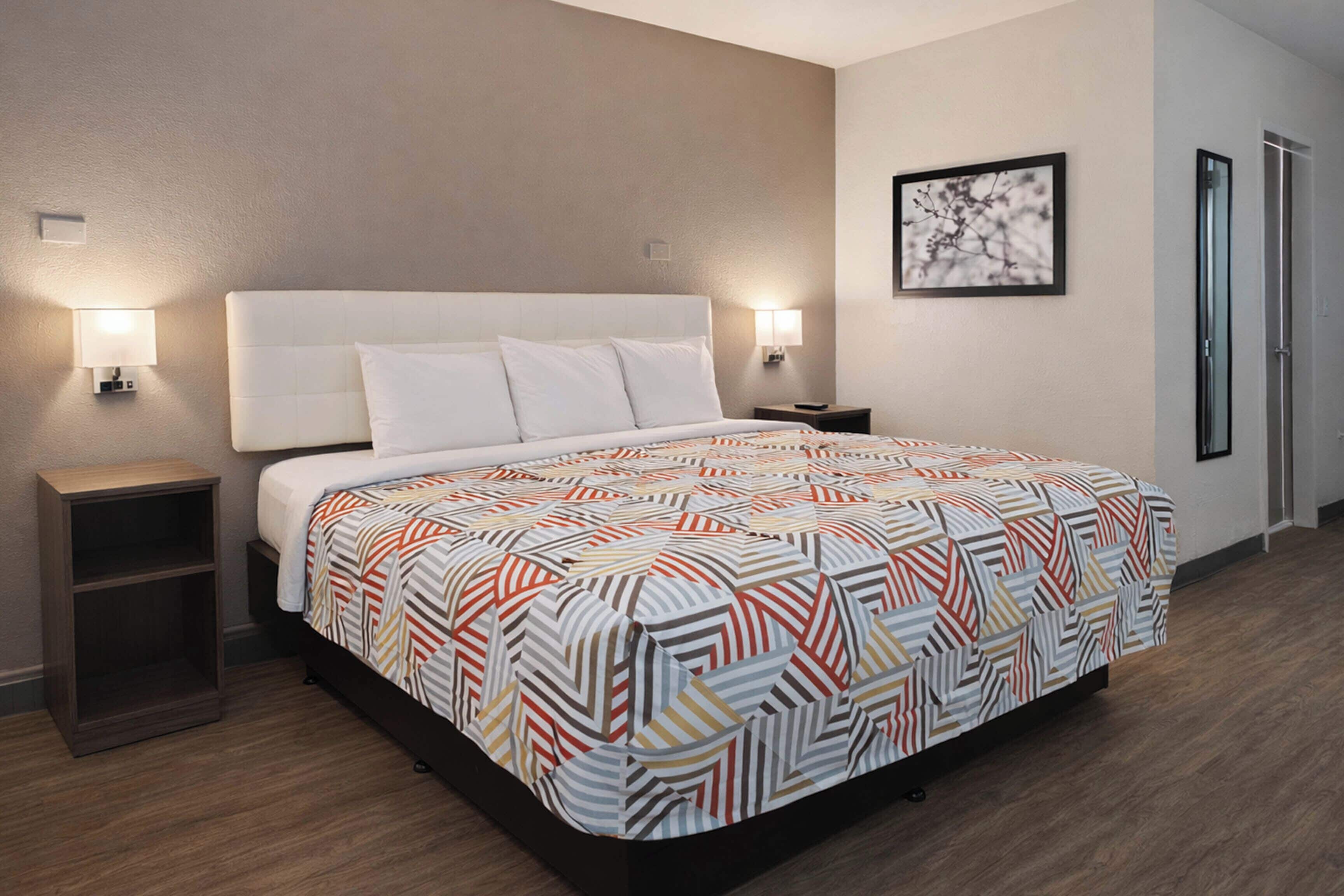 Quarto de hotel com uma cama grande, duas luminárias de cabeceira, uma mesa de cabeceira, uma mesinha de madeira e um quadro na parede.