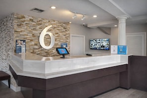 A reception desk with a large number '6' sign, a mounted television, and a digital information kiosk. - Motel 6 – Lake City, Fl – Medical Center (Lake City)