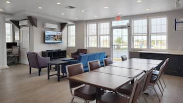 A spacious room with a large table and chairs, a television mounted on the wall, and large windows providing natural light.