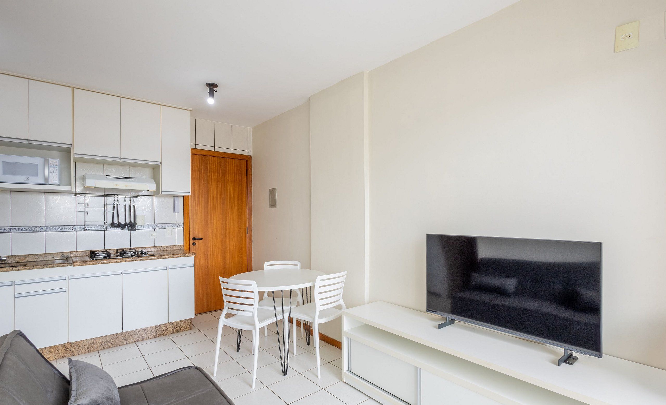 Een moderne keuken met witte kasten, een magnetron en een fornuis. Er is een eethoek met een witte tafel en stoelen, en een flatscreen televisie bevestigd aan een witte kast.
